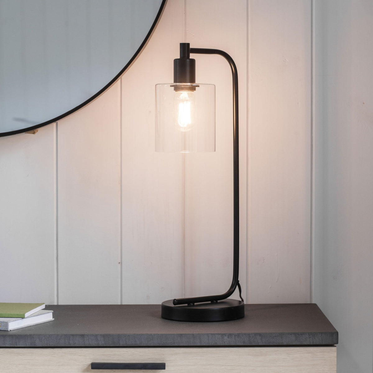 Black metal table lamp with clear glass shade and exposed bulb on a wooden dresser with a round mirror above.