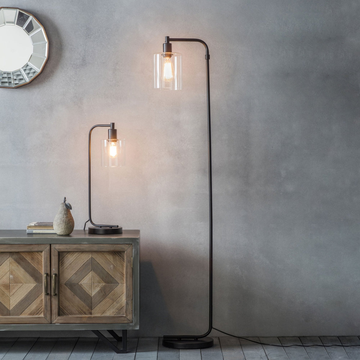 Matching black metal floor and table lamps with clear glass shades next to a wooden cabinet and a round mirror.