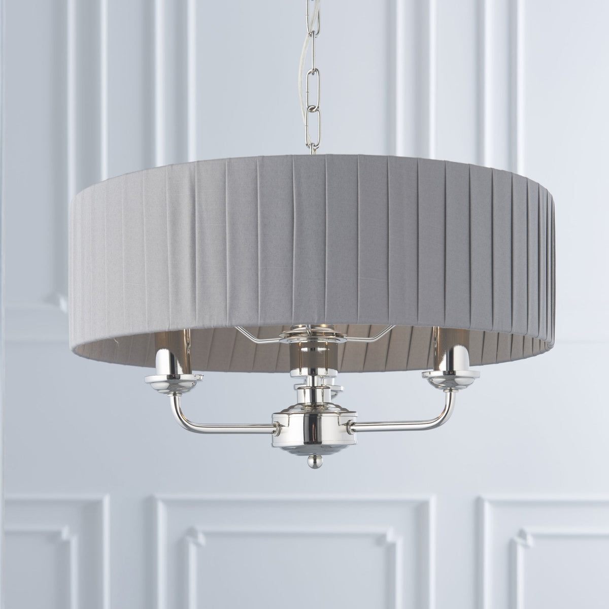 A grey pleated drum shade chandelier with three lights hangs against a white paneled wall.