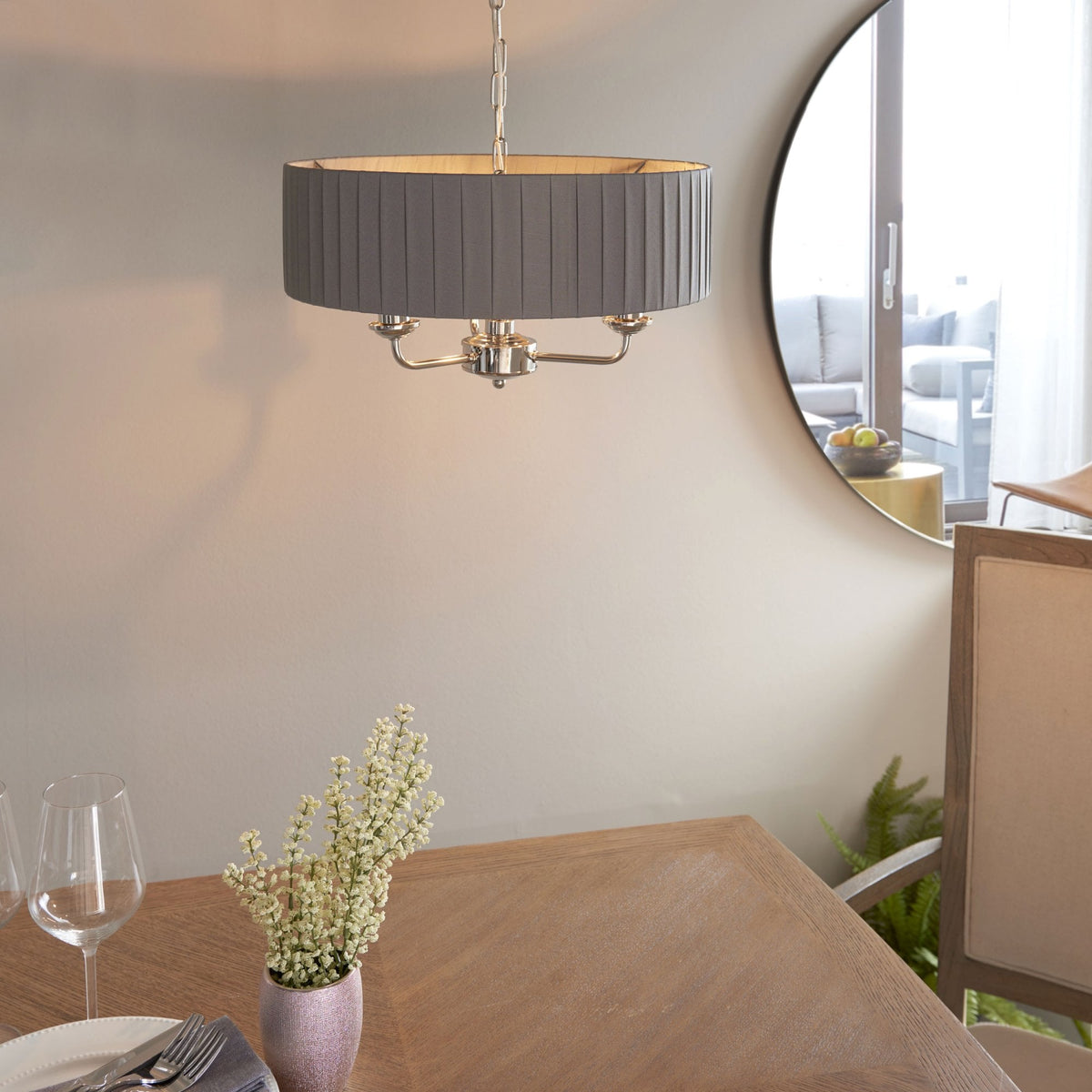 A grey pleated drum shade chandelier hangs above a dining table with a vase of flowers and a round mirror.