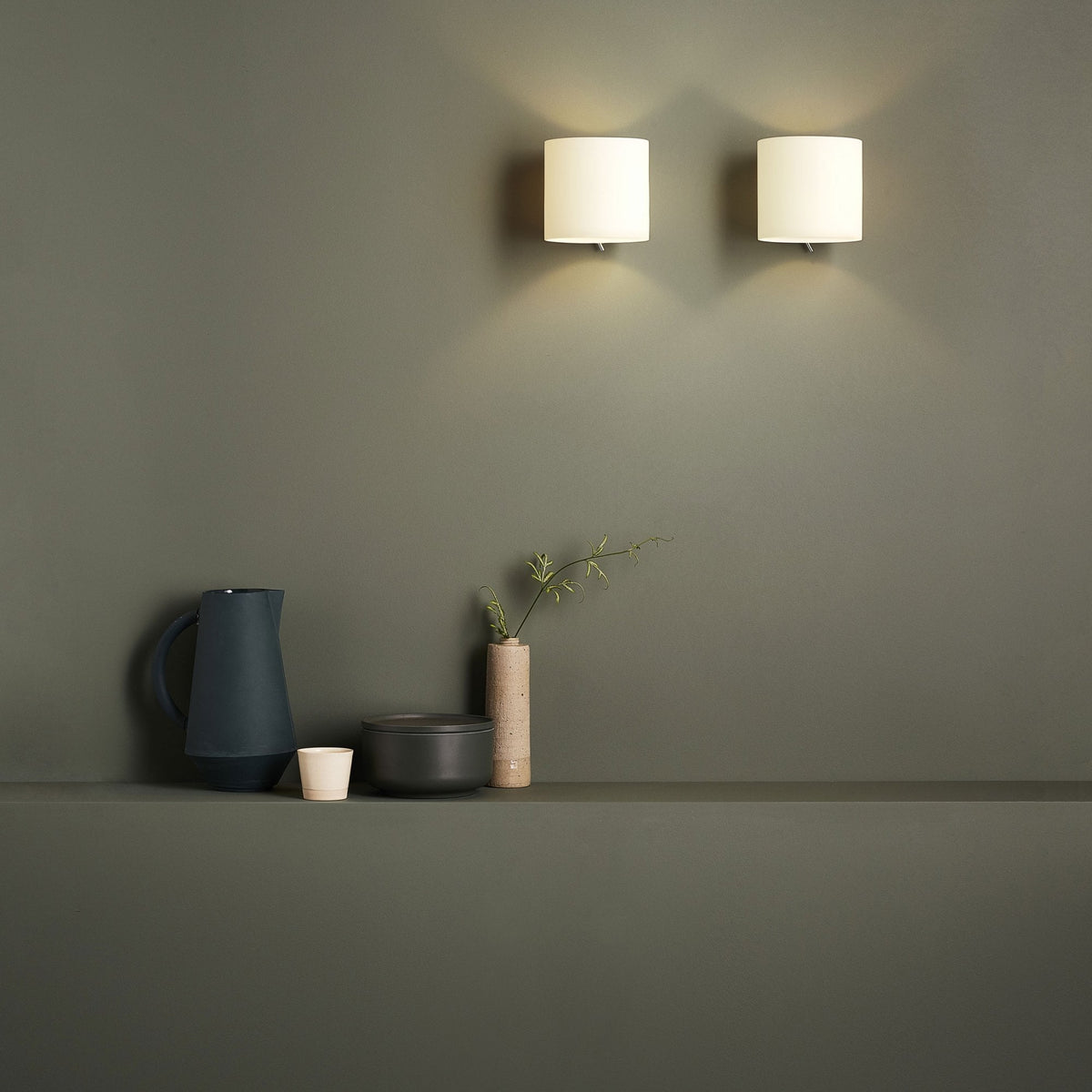 Two wall sconces illuminate a dark green wall above a shelf with a pitcher, cup, lidded bowl, and vase