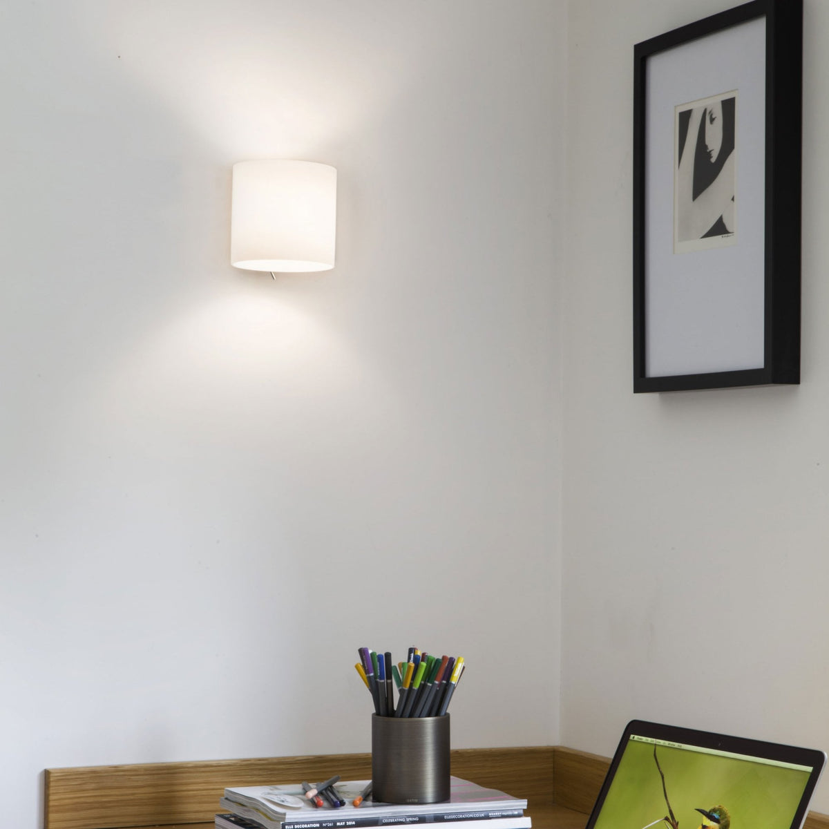 White cylindrical wall light illuminating a white wall above a wooden desk with a laptop and pens