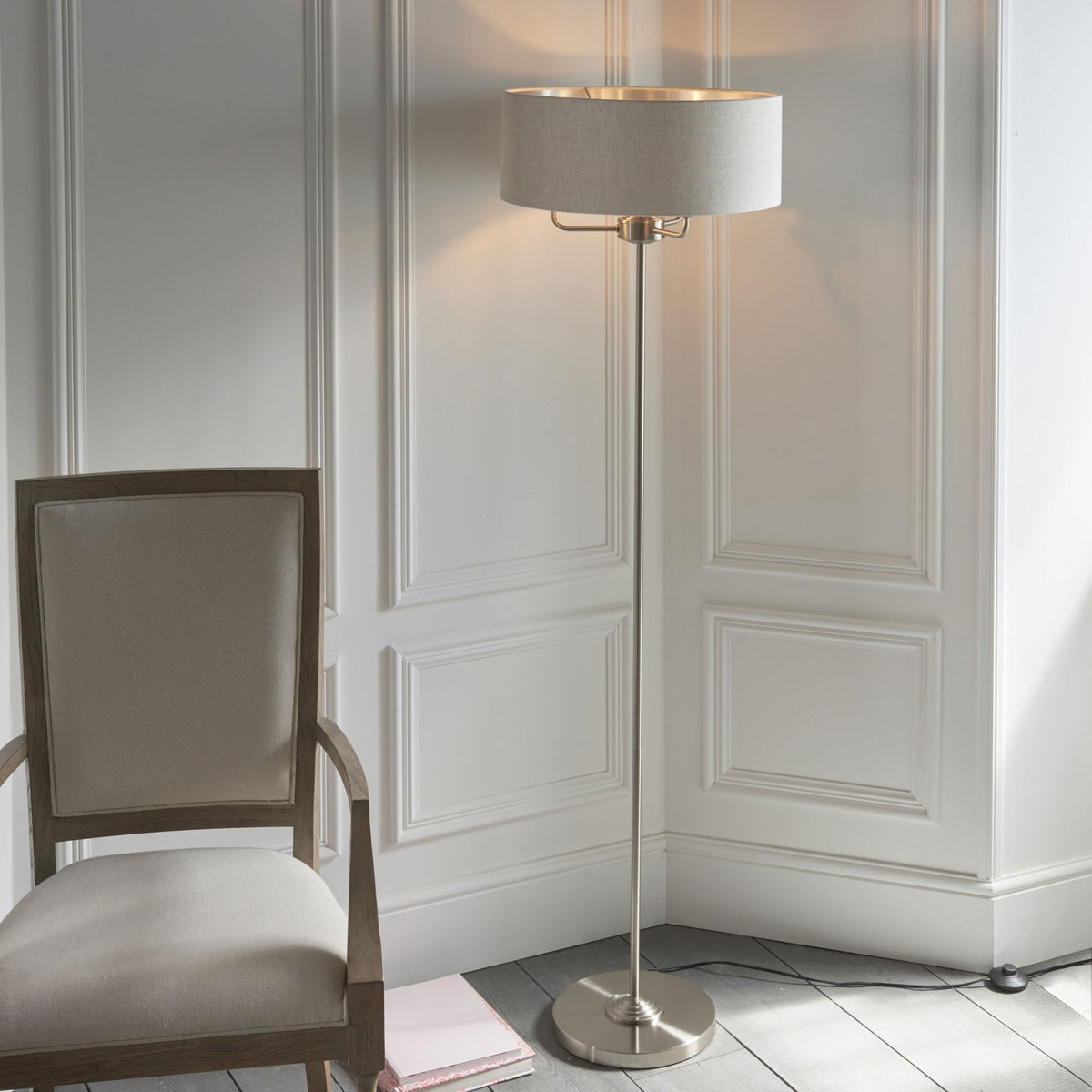 A brushed nickel floor lamp with a light fabric shade stands next to a light-colored chair.