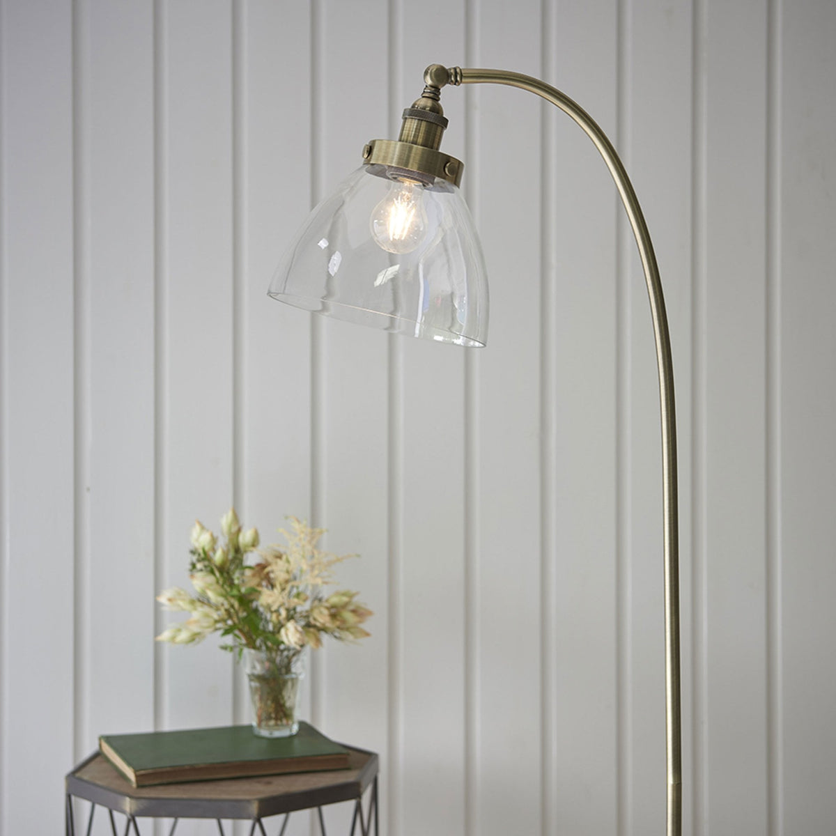 Antique brass floor lamp with a clear glass shade, illuminating a small bouquet of flowers on a side table.