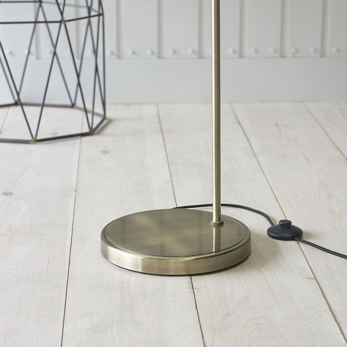 Close-up of the round, antique brass base of a floor lamp on a light wooden floor, with a black power cord.