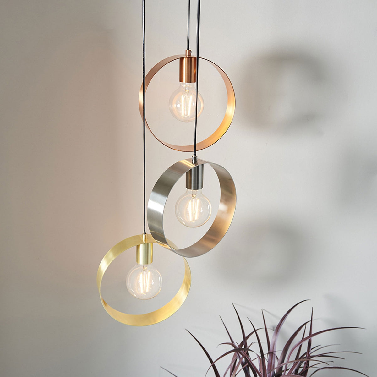 Three pendant lights with circular metal shades in copper, silver, and gold, illuminating a room with a plant in the foreground.