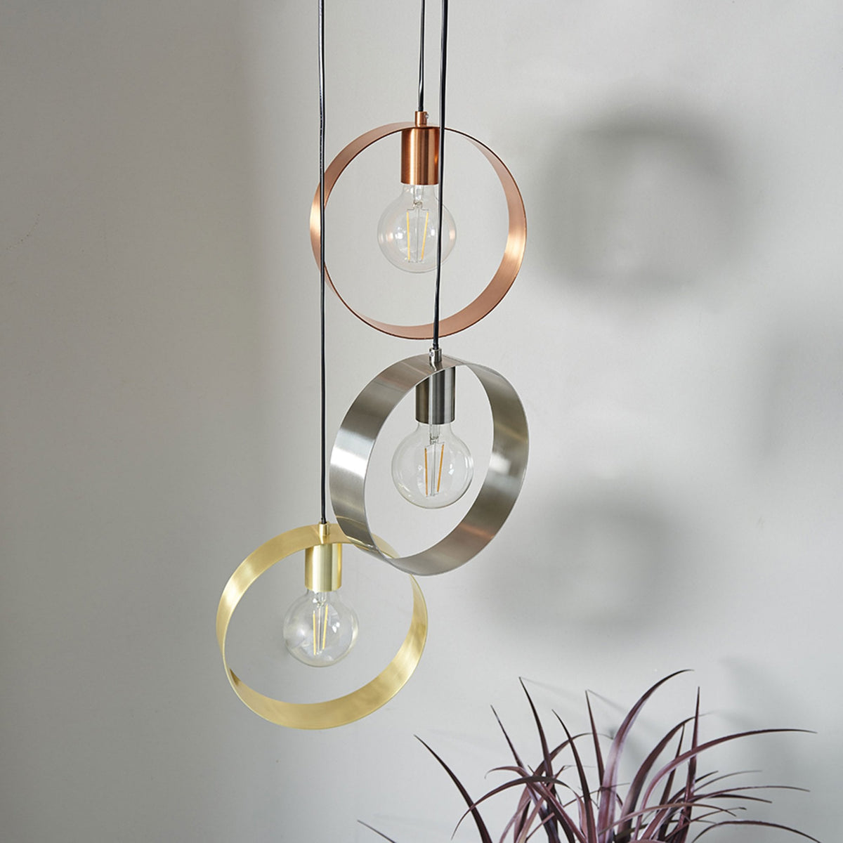 Three pendant lights with circular metal shades in copper, silver, and gold, illuminating a room with a plant in the foreground.
