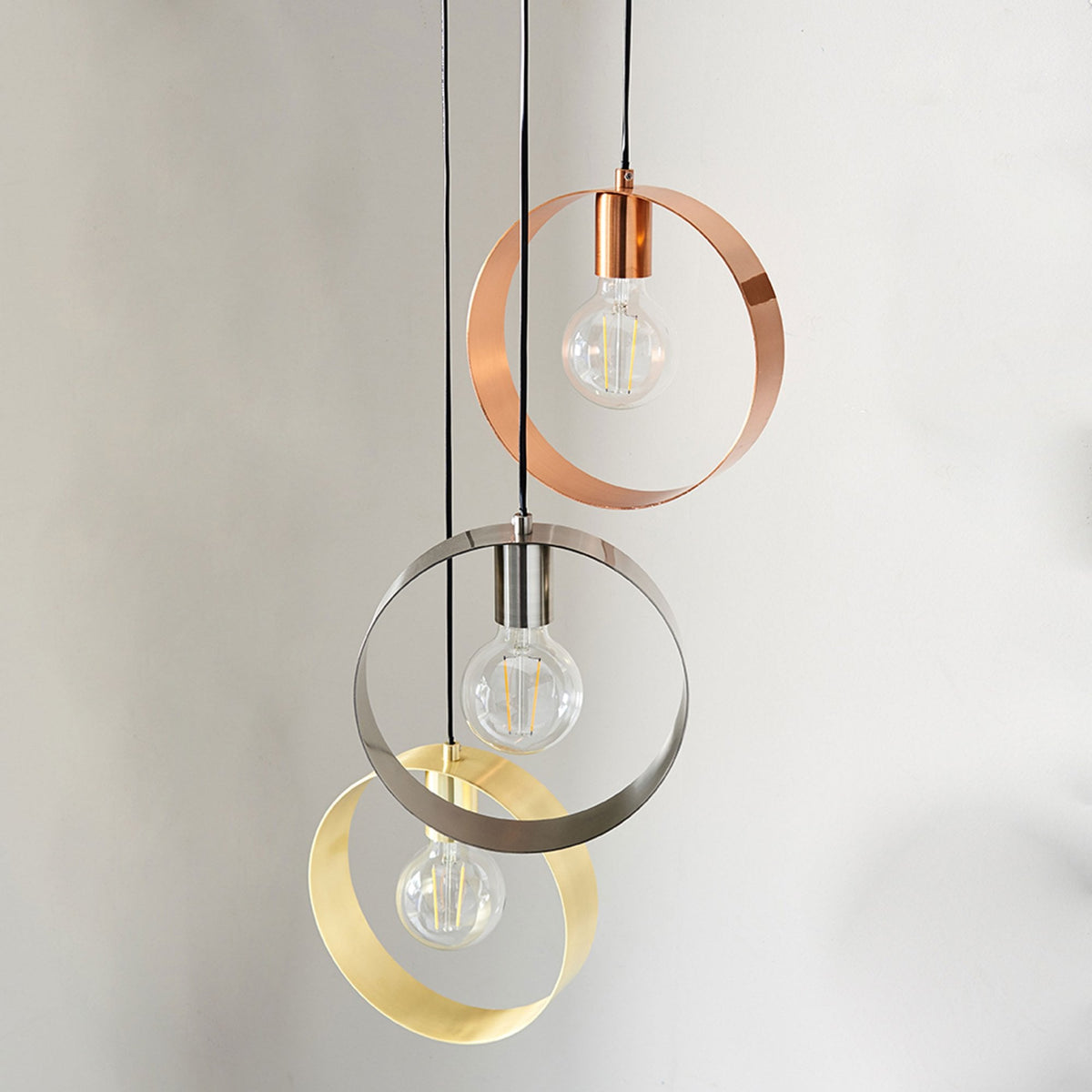 Three pendant lights with circular metal shades in copper, silver, and gold, illuminating a room with a plant in the foreground.