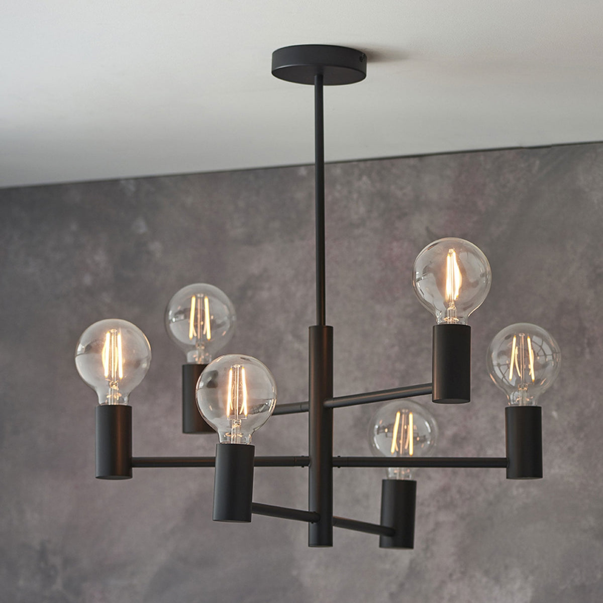 A black chandelier with six exposed bulbs hangs from a gray ceiling against a dark gray wall.