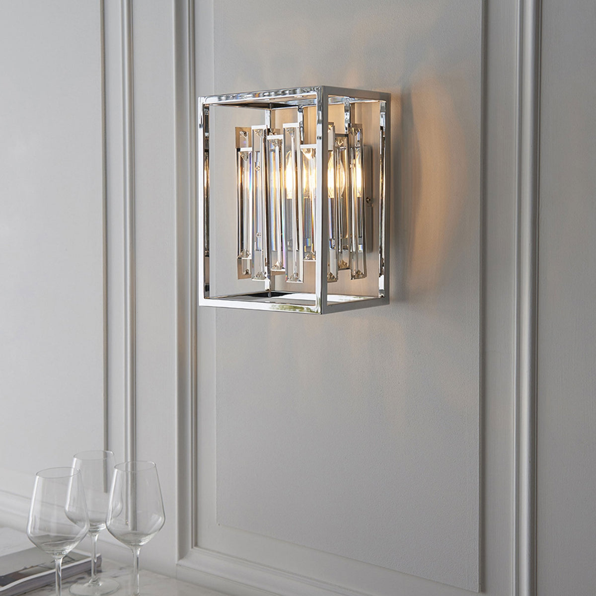 Chrome and crystal wall light illuminates a white paneled wall above two wine glasses.