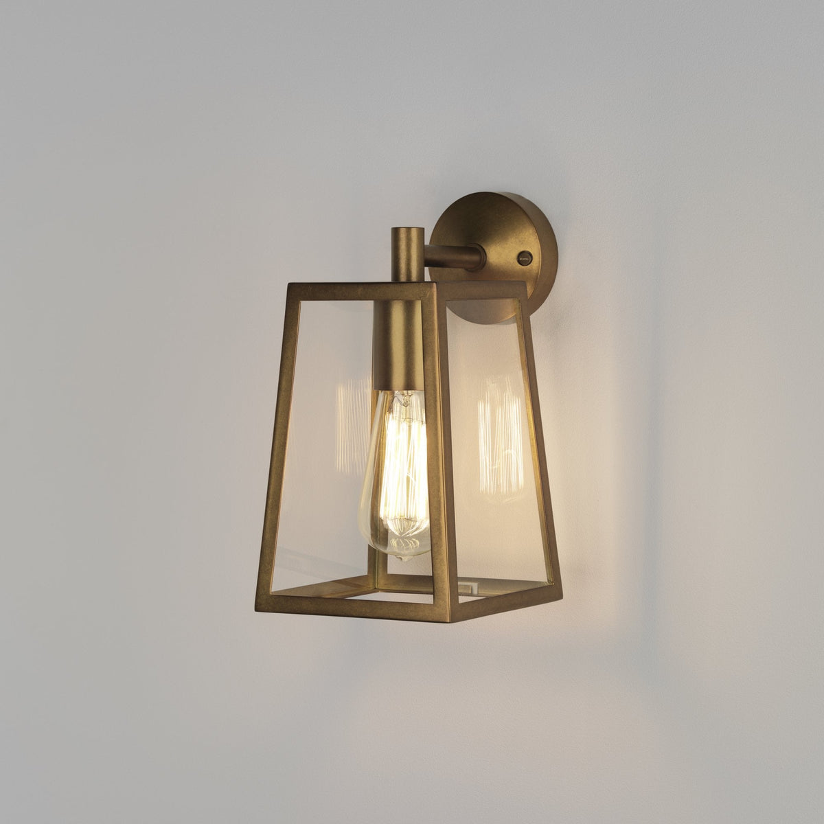 A brass wall light with a clear glass shade and an exposed filament bulb