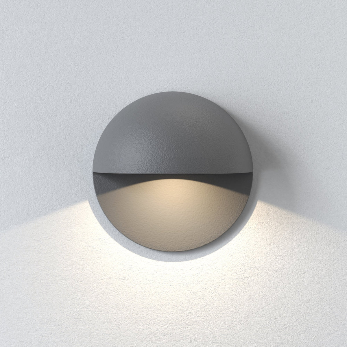 Round grey wall light with a textured finish, emitting a warm glow downwards onto a white textured wall