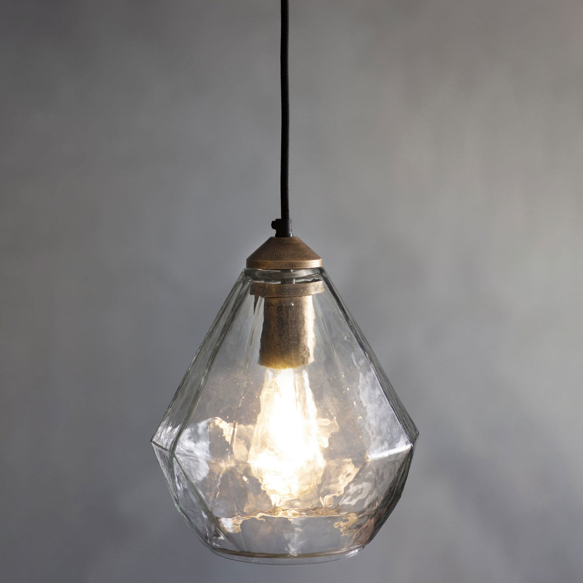 Clear glass pendant light with a faceted, diamond-like shape and a visible filament bulb, hanging against a gray wall.