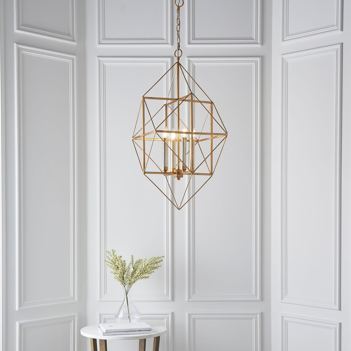 A gold geometric chandelier with four lights hangs in a stylish room with white wainscoting and a small table.