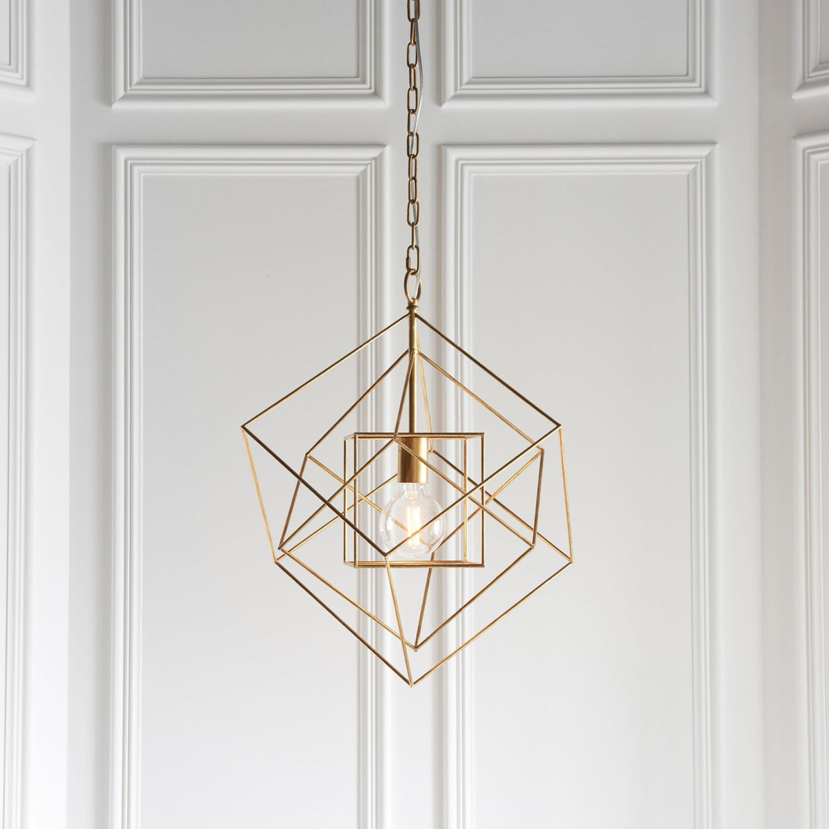 A brass geometric pendant light with an exposed bulb hanging against a white paneled wall.