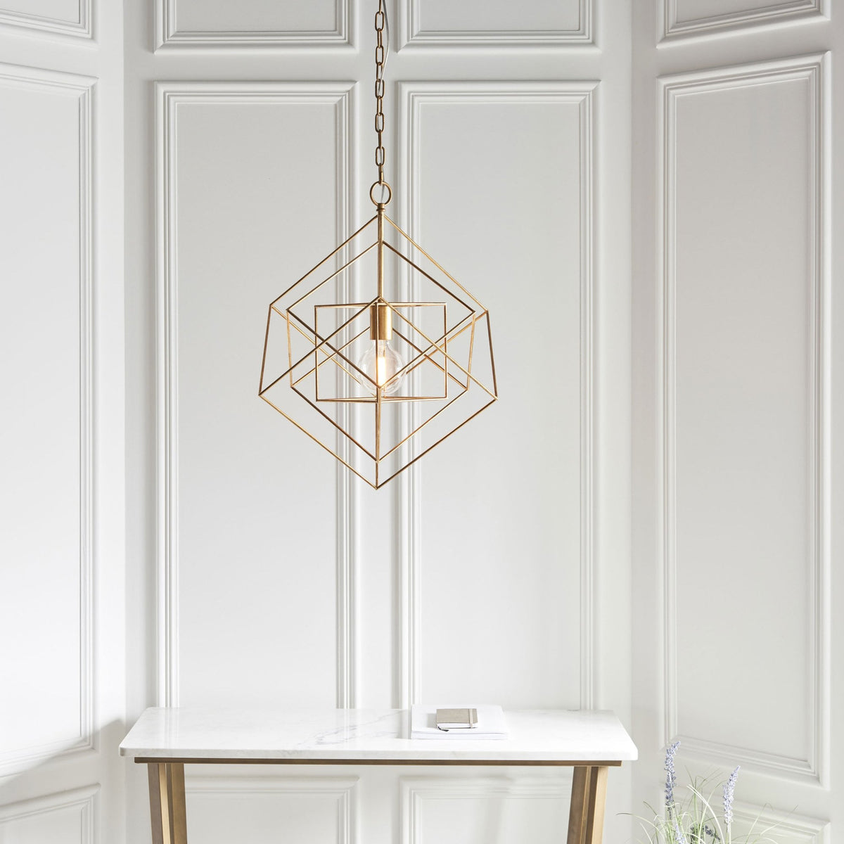 A brass geometric pendant light hanging above a minimalist console table against a white paneled wall.
