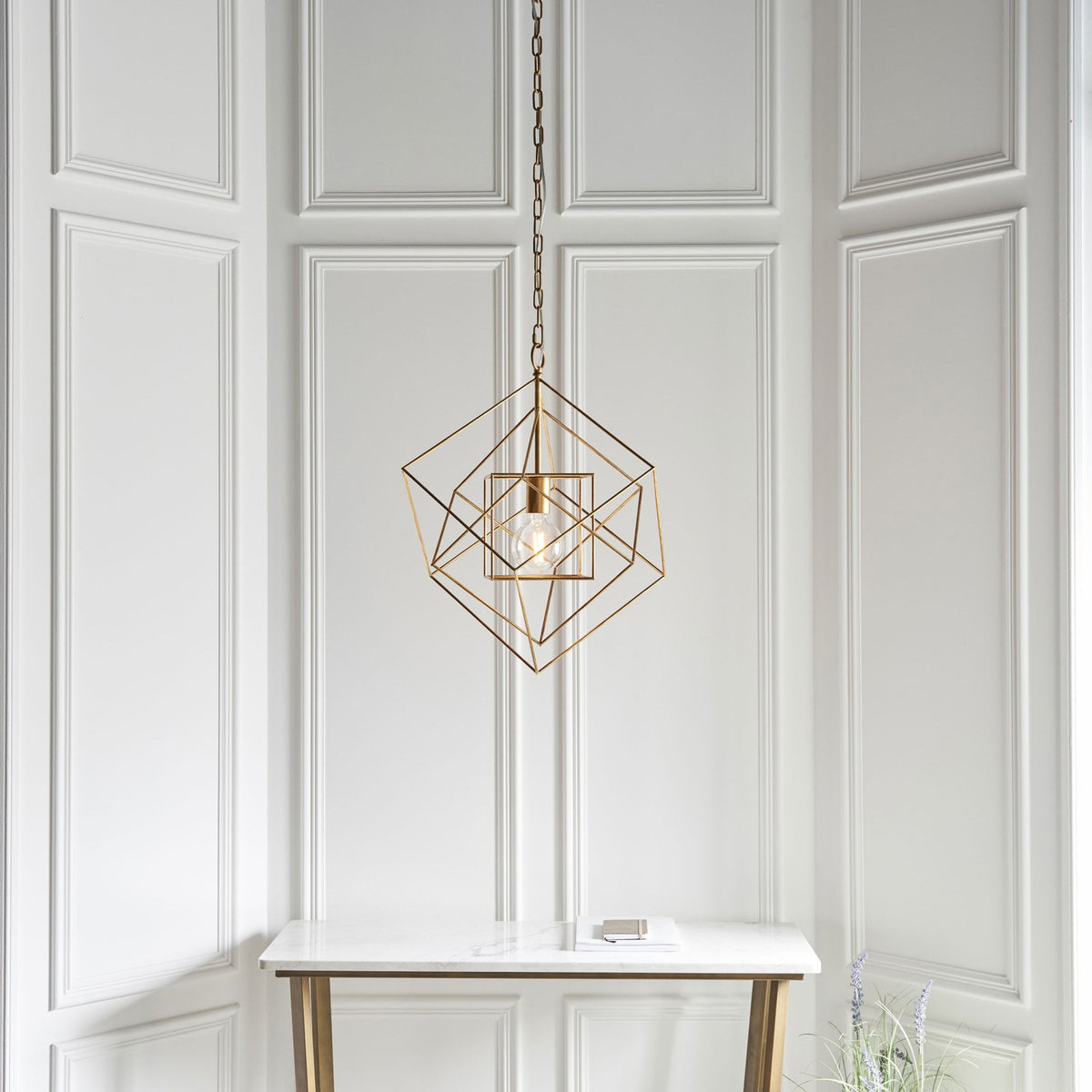 A brass geometric pendant light above a simple white console table in a room with white paneled walls.