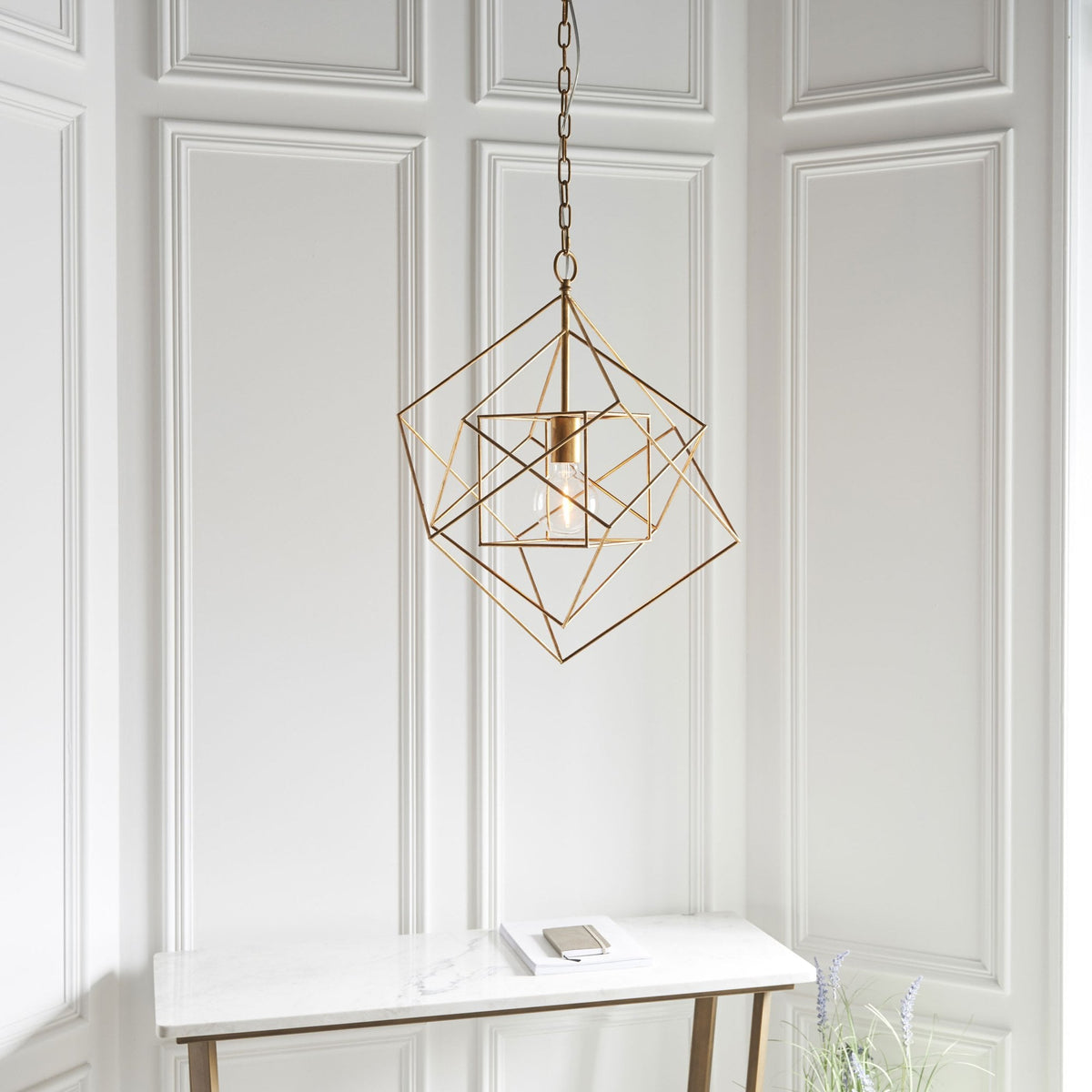 A brass geometric pendant light illuminating a white console table in a room with wainscoting.