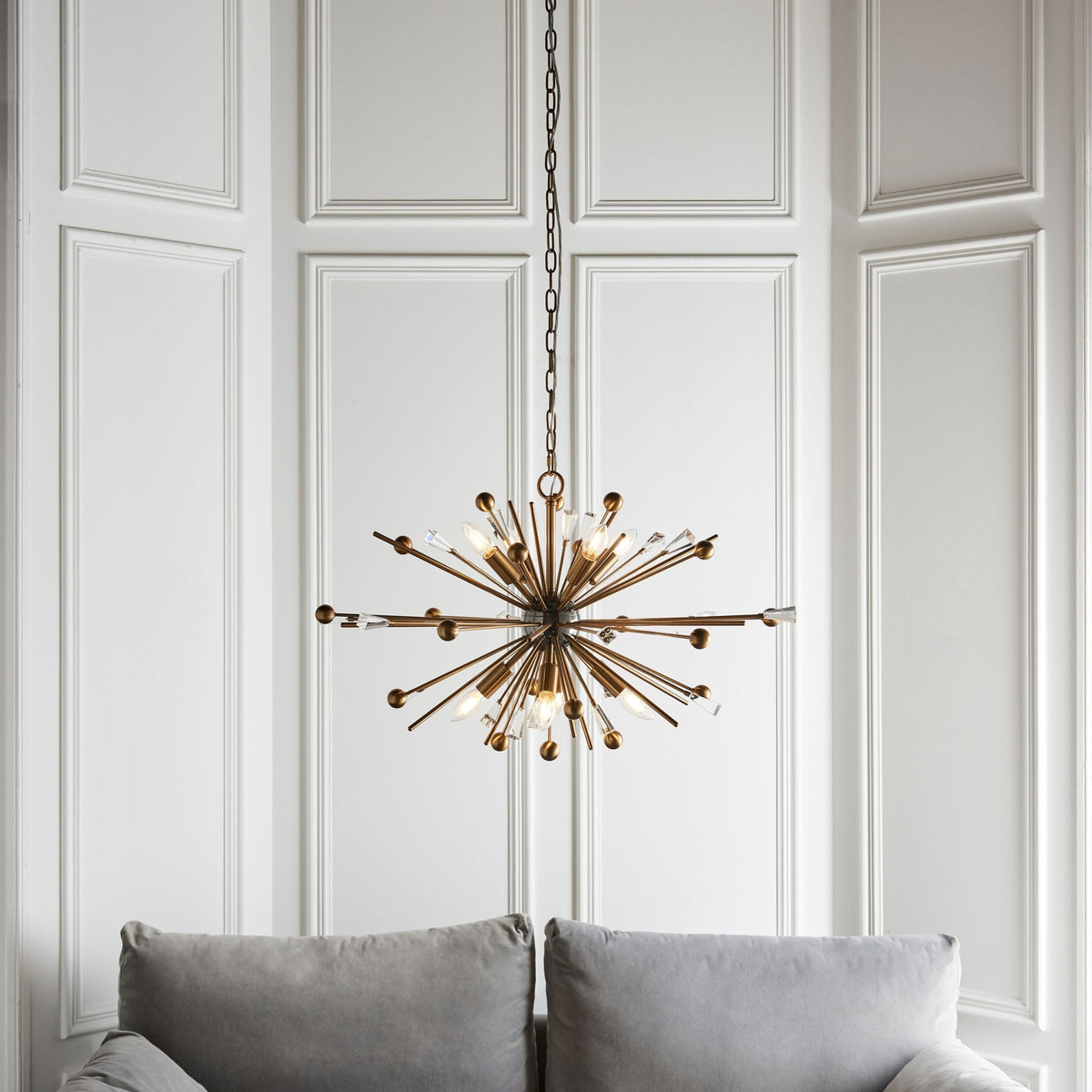 A sputnik-style chandelier with brass rods and crystal accents hangs above a gray sofa against a paneled wall.