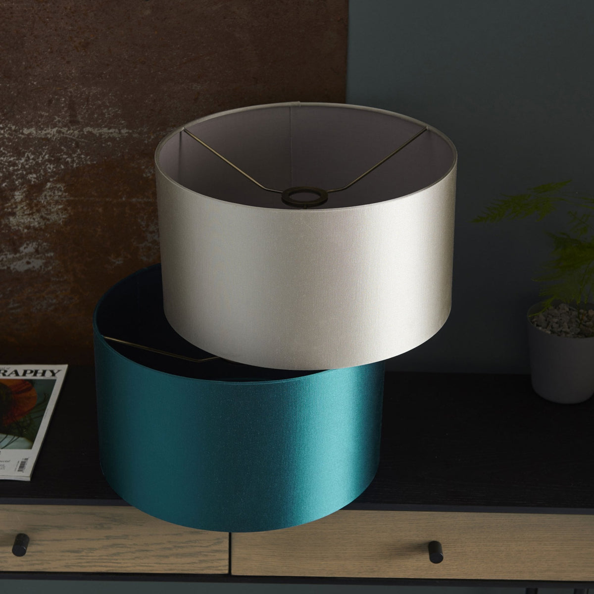 Two drum lampshades, one teal and one cream, are stacked on a wooden dresser in a contemporary setting.