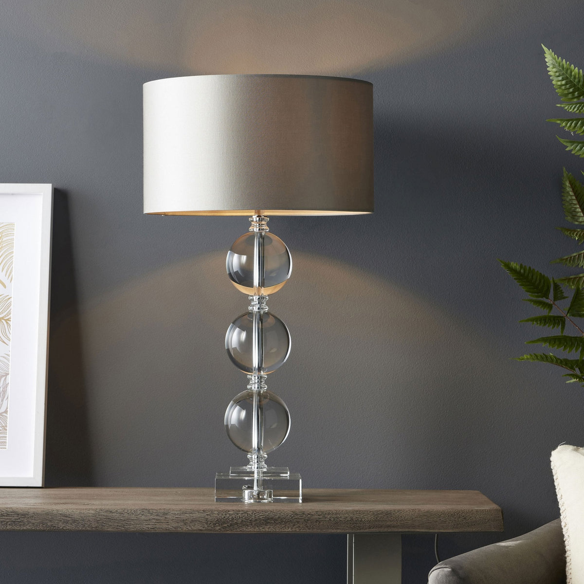 A table lamp with a clear glass base featuring three stacked spheres and a light grey drum shade, casting a warm glow.