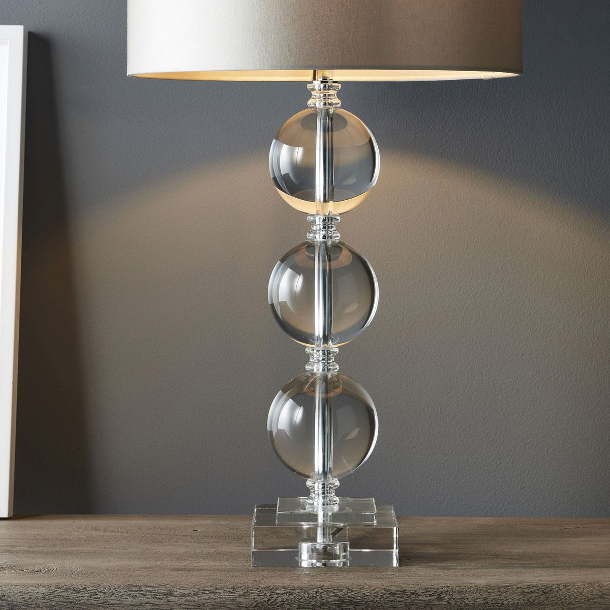 Close-up of a table lamp with a clear glass base featuring three stacked spheres and a light grey drum shade.