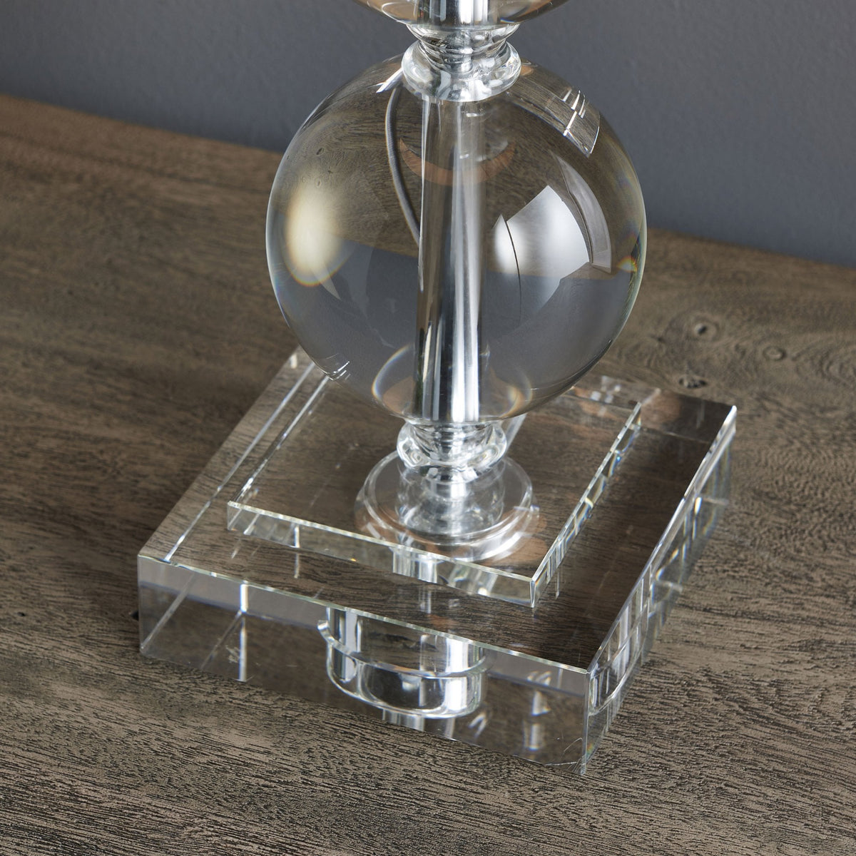 Close-up of the clear glass square base and bottom sphere of a table lamp on a wooden surface.