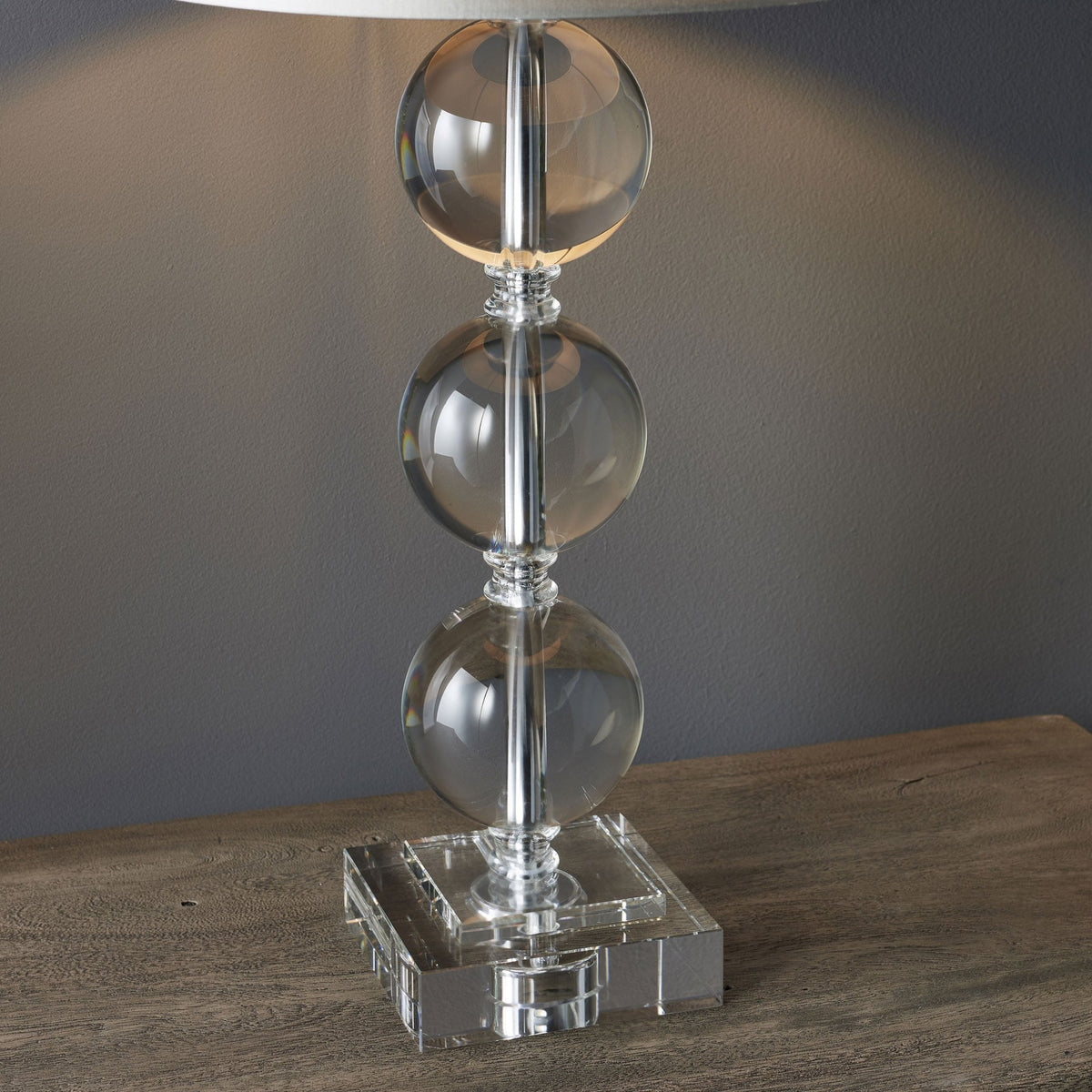 A table lamp with a clear glass base featuring three stacked spheres and a light grey drum shade, casting a warm glow.