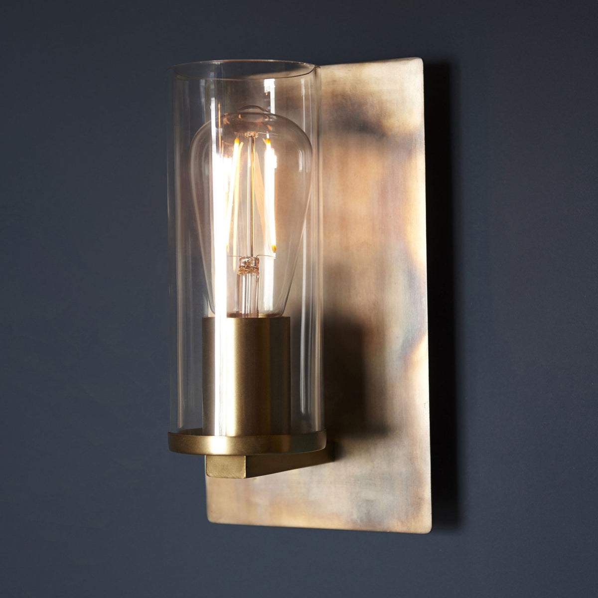 A close-up of a wall light, featuring a clear glass cylinder and a brass base.