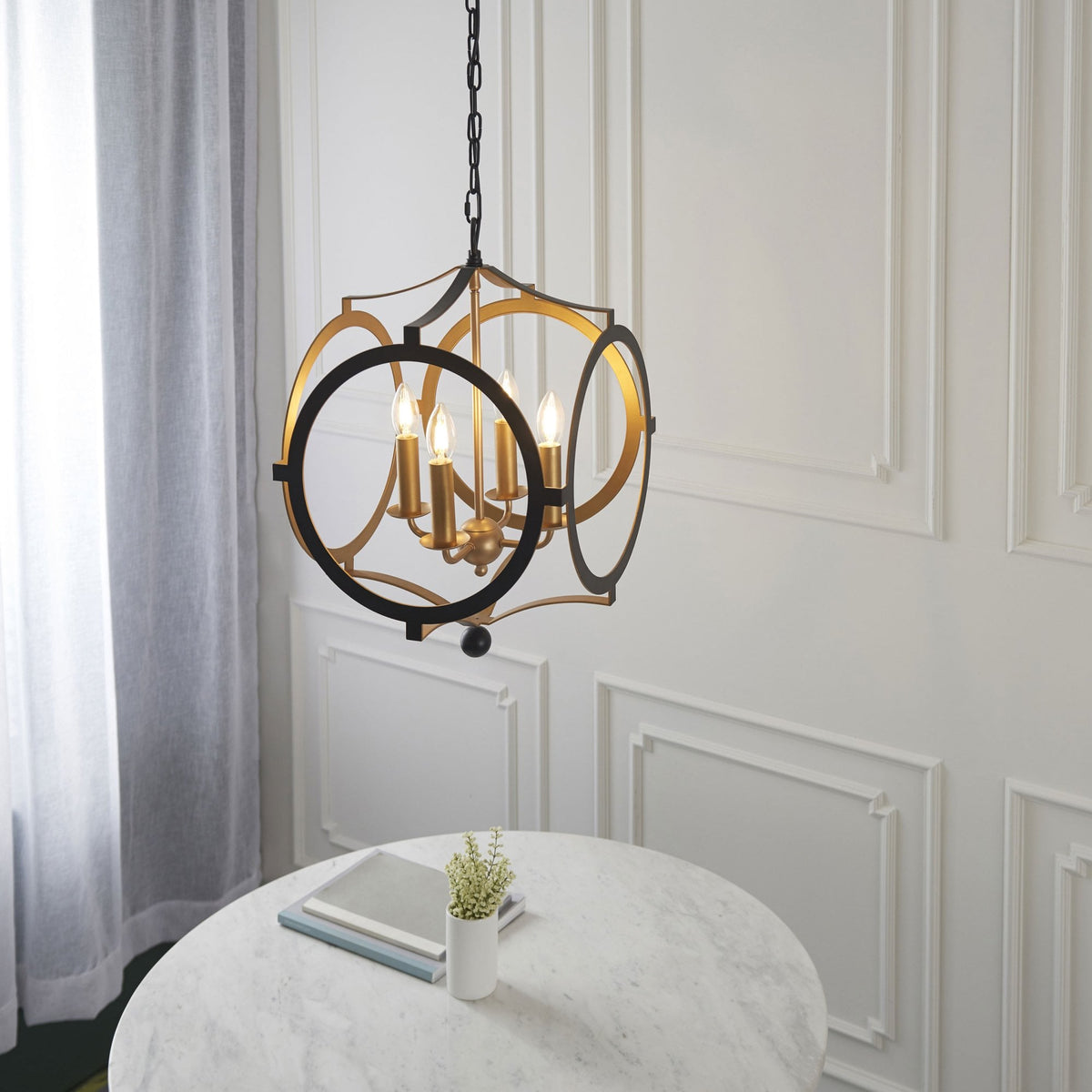 A black and gold chandelier hangs above a white marble table with a book and plant.
