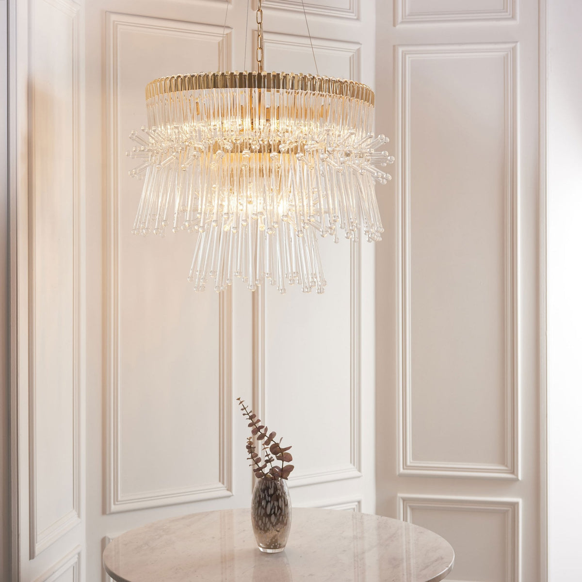 Two-tier gold and clear glass chandelier hanging above a round table with a vase of dried flowers.