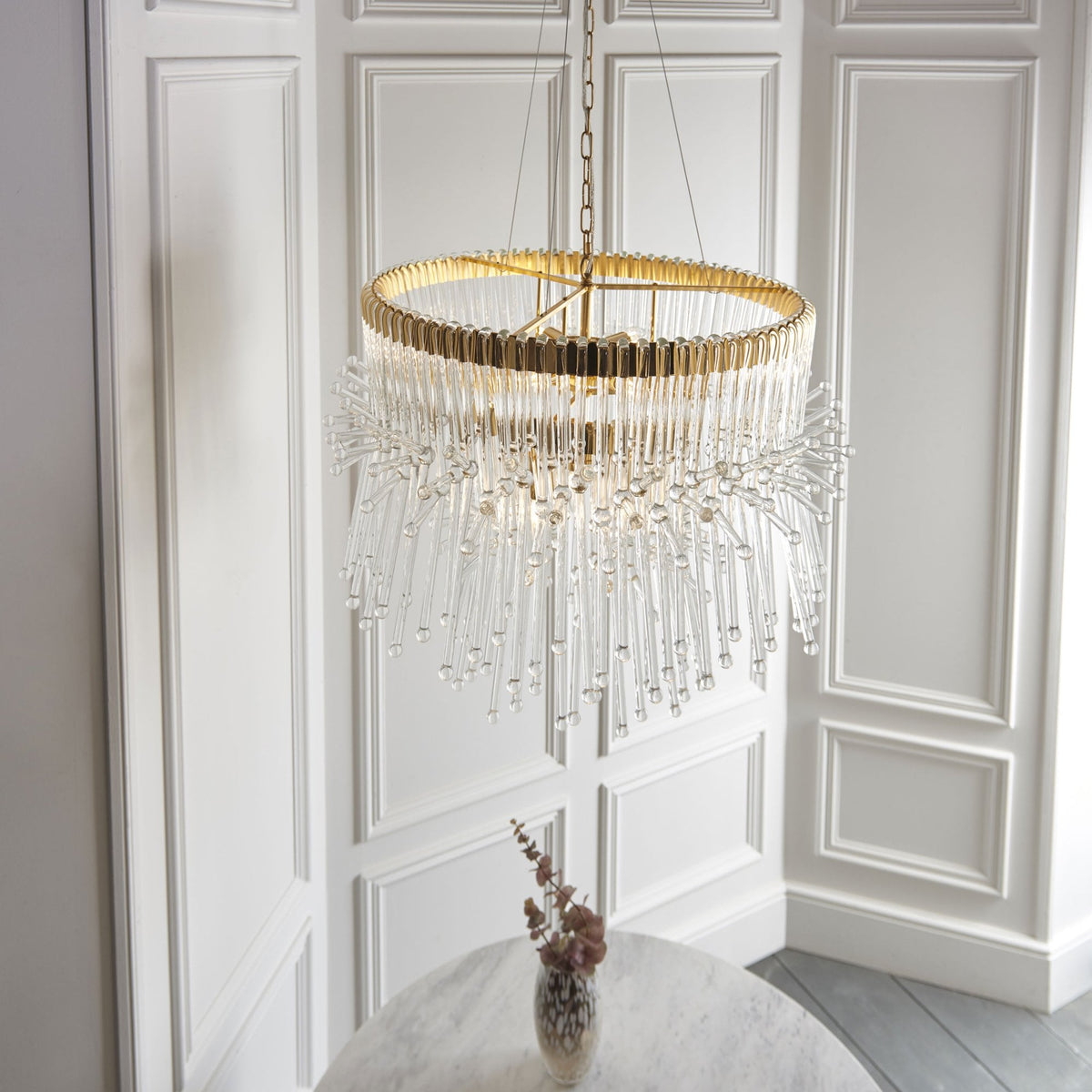 Two-tier gold and clear glass chandelier illuminating a white room with a round table and vase.