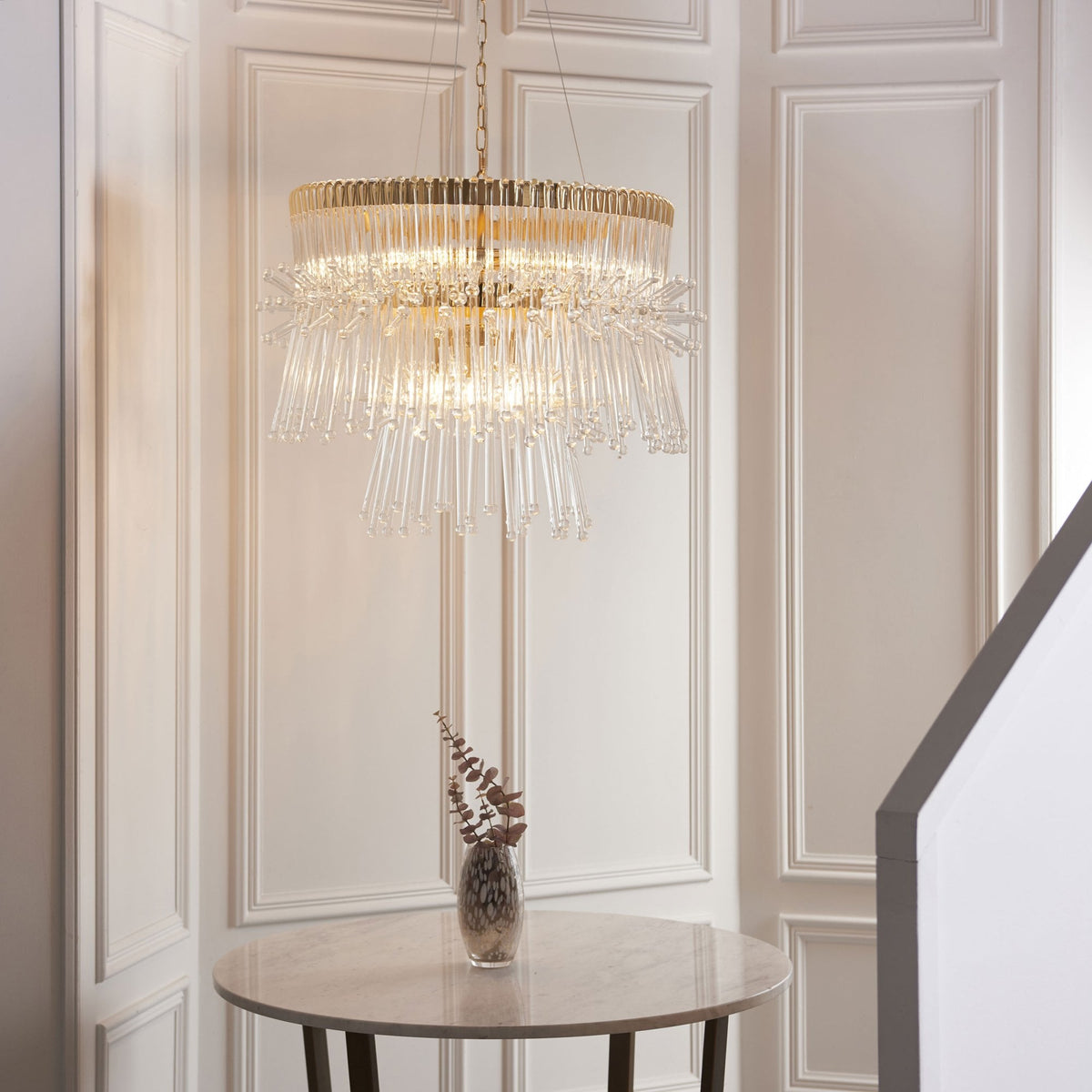 Gold and clear glass chandelier above a round table with a vase, in a room with white paneled walls.