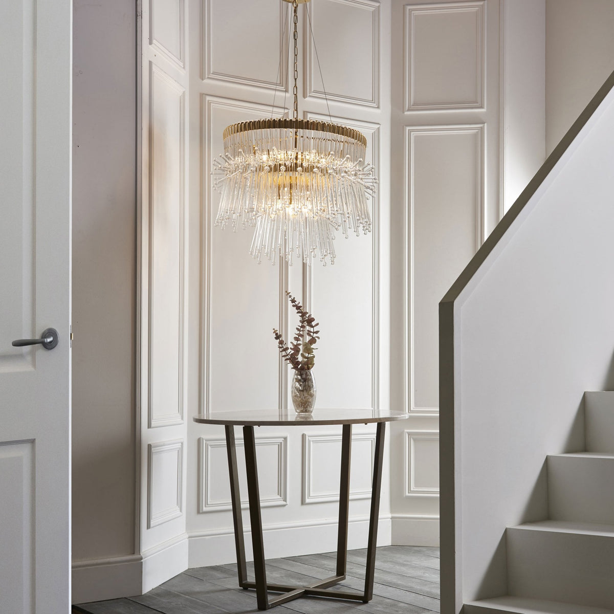 Gold and clear glass chandelier in a hallway with white paneled walls, a round table, and stairs.