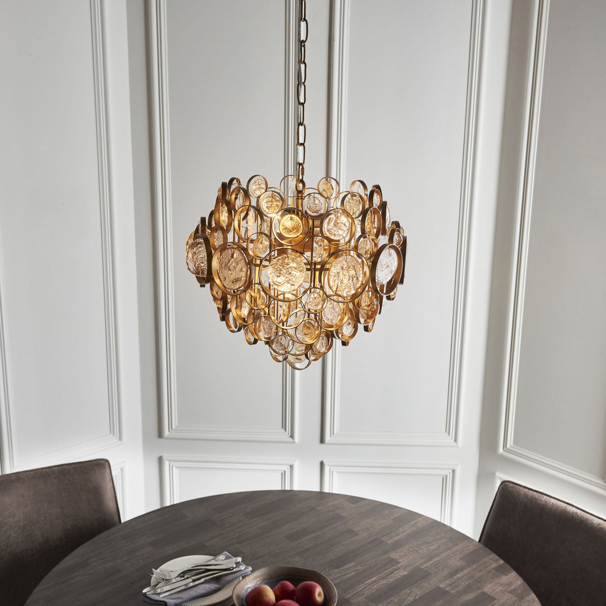 A gold chandelier hangs above a dark round dining table with two chairs, set against white paneled walls.