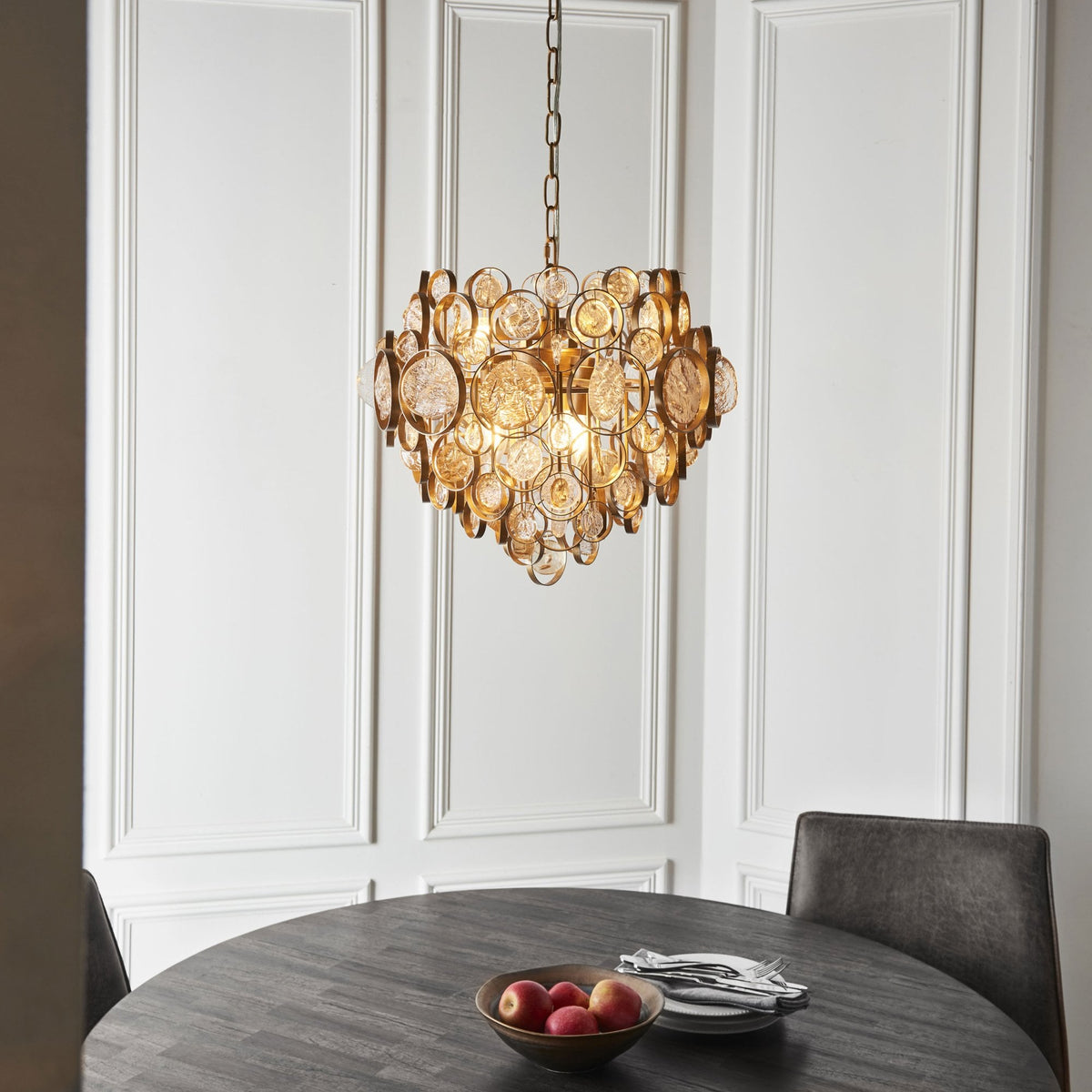 Elegant gold chandelier suspended above a dark round dining table and two chairs in a brightly lit room.