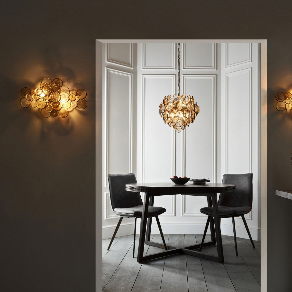 A gold chandelier is centered above a dark dining table with two chairs in a chic room with sconces.