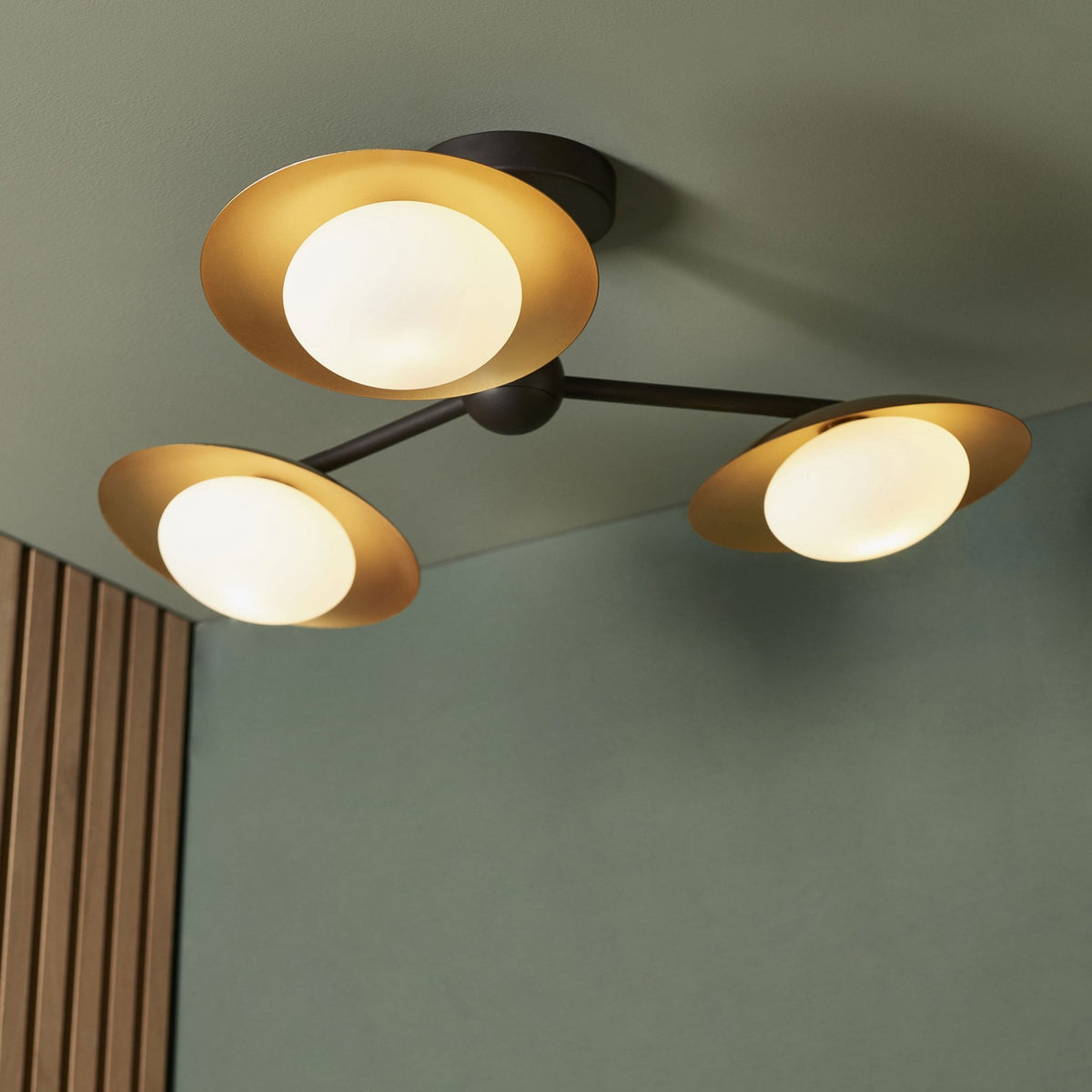 A stylish ceiling light with three gold-shaded spherical bulbs, casting a warm glow on a green wall.