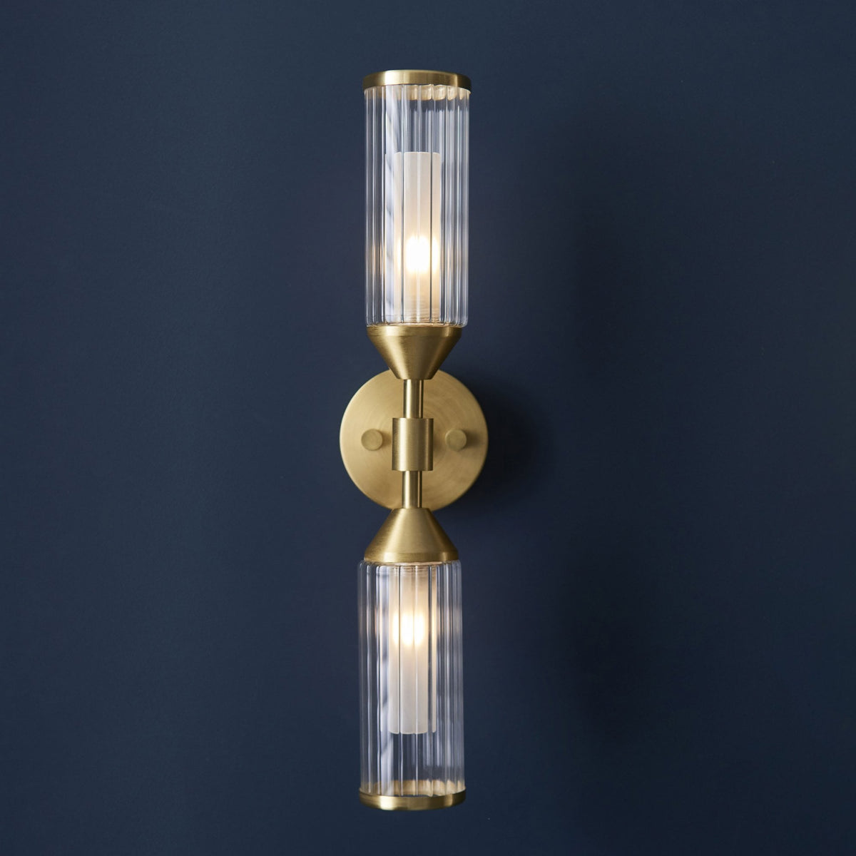 A brass wall light with two clear, fluted glass shades illuminating a dark blue wall.