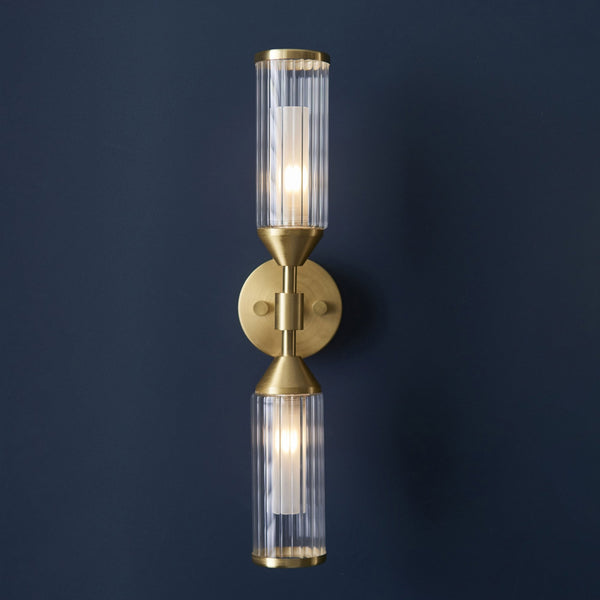 A brass wall light with two clear, fluted glass shades illuminating a dark blue wall.