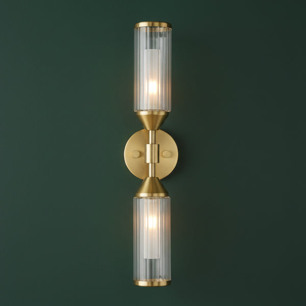 A brass wall sconce with two lit ribbed glass shades, one pointing up and one down, on a white background.