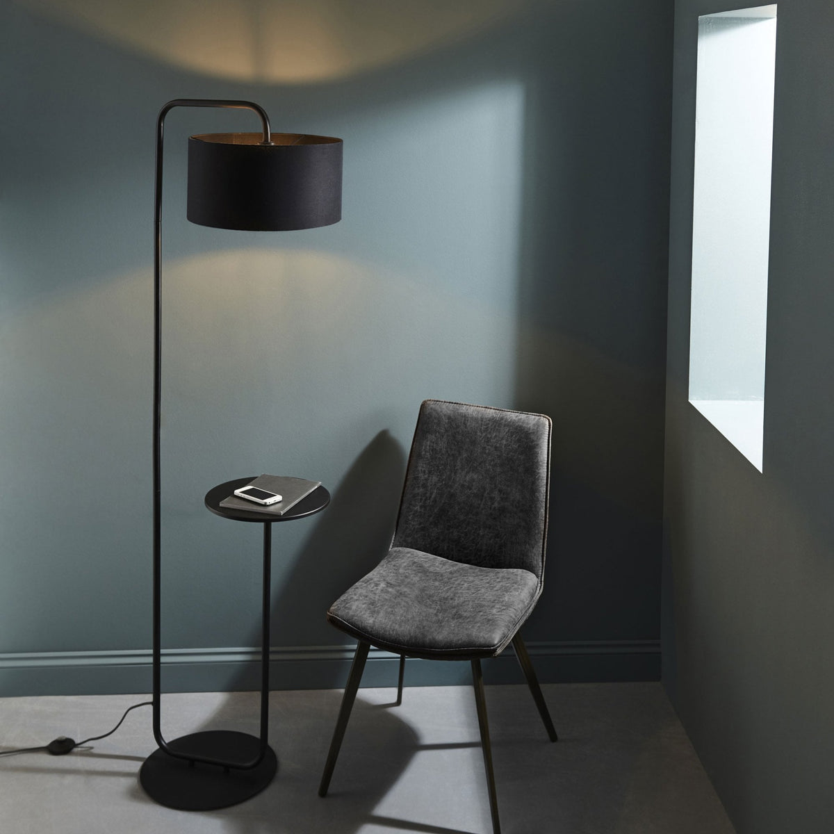 Black floor lamp with a round side table attached, next to a grey upholstered chair.