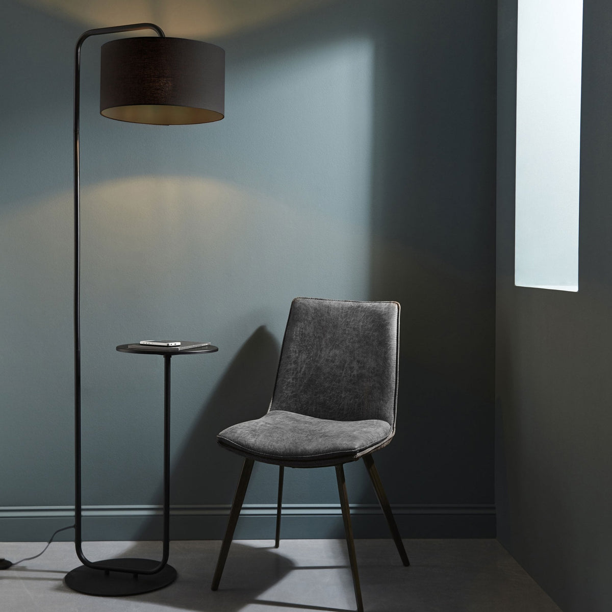 Black floor lamp with a round side table attached, next to a grey upholstered chair in a room.