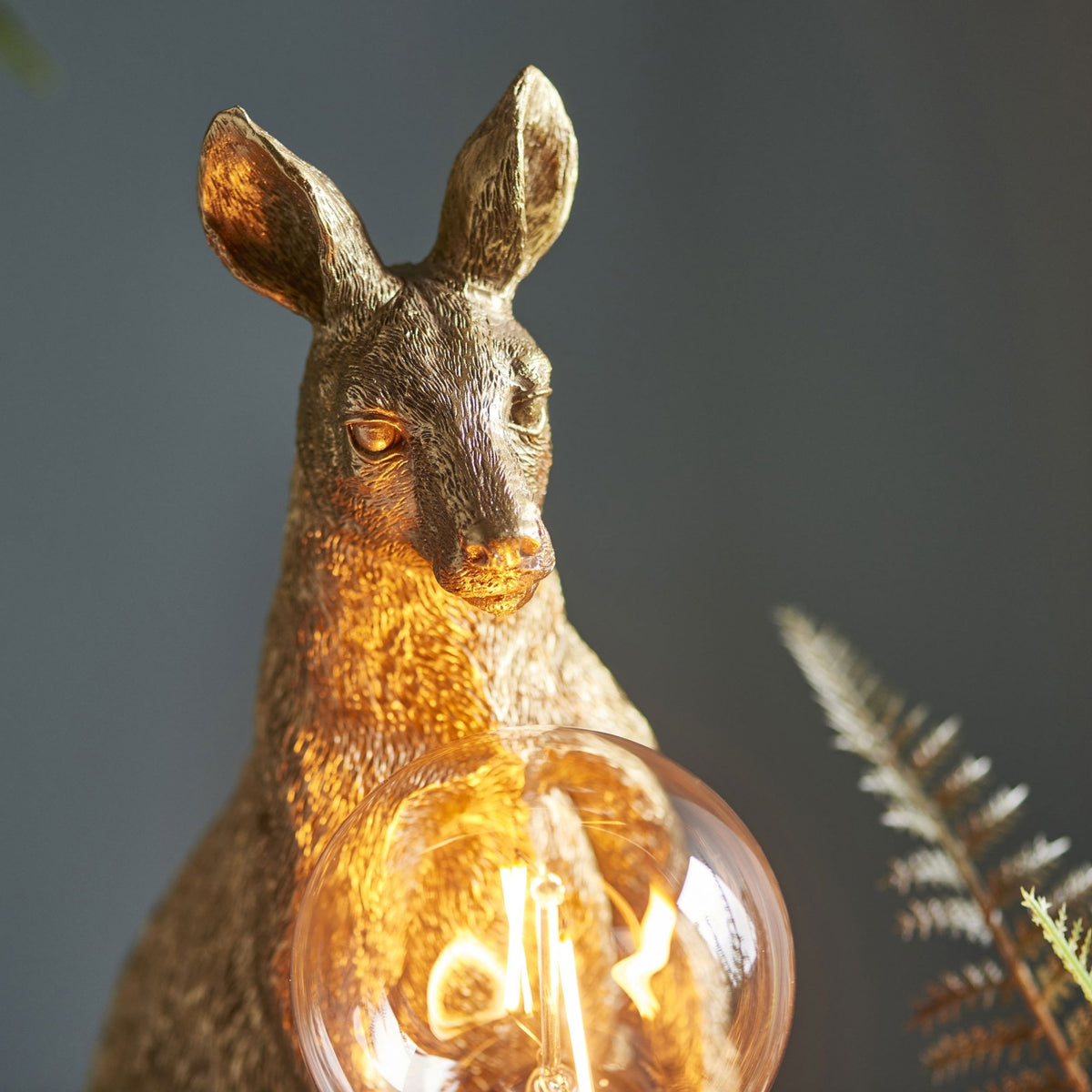 Golden kangaroo lamp with a glowing bulb, set against a dark background with subtle foliage.