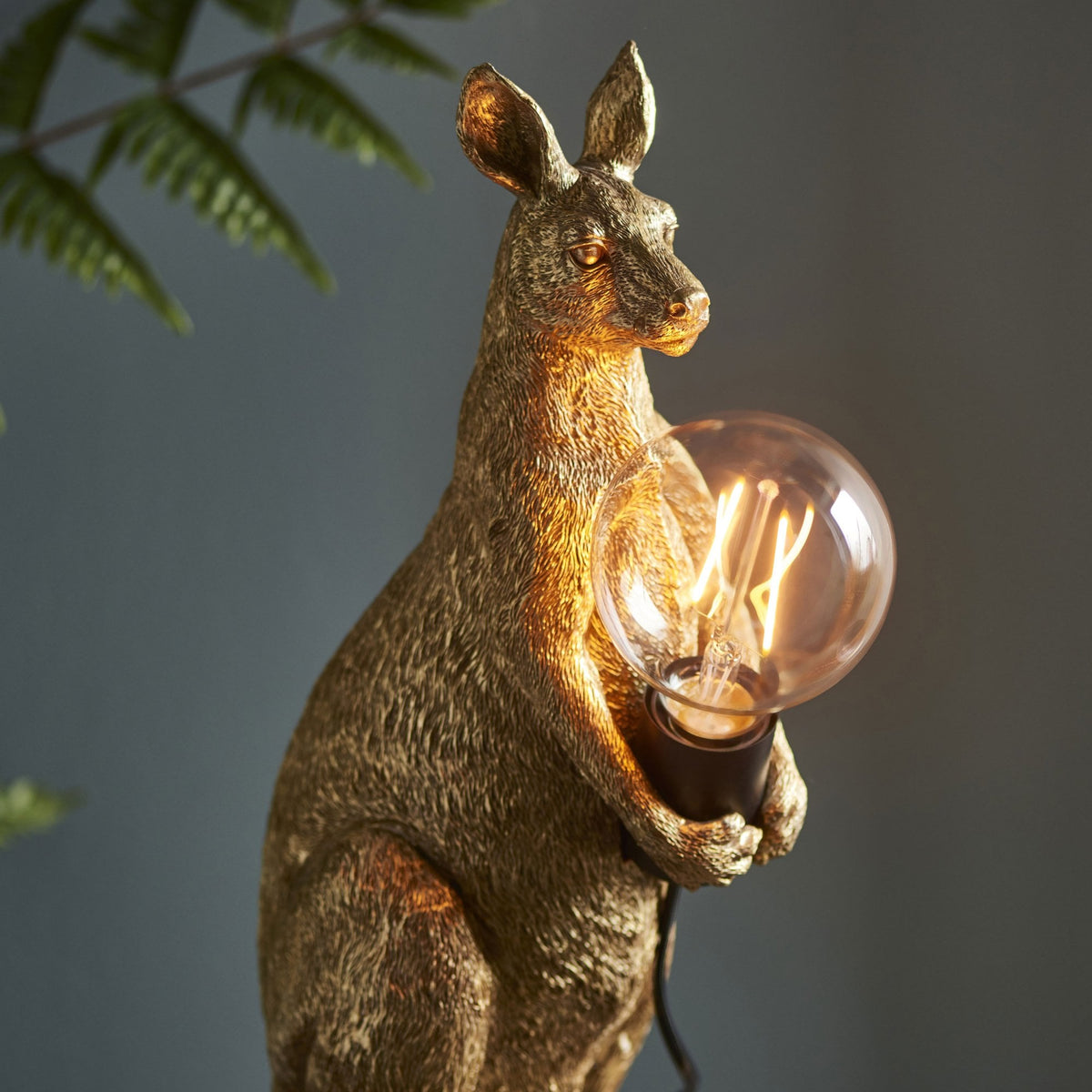 Golden kangaroo table lamp holding a glowing bulb, with lush green plants in the background.