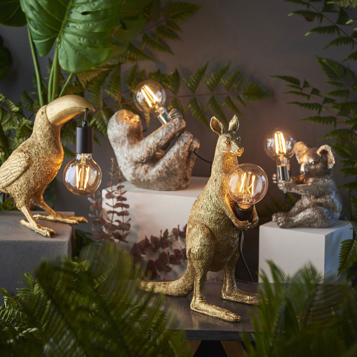 Collection of golden animal lamps: toucan, sloth, kangaroo, and koala, amidst tropical foliage.