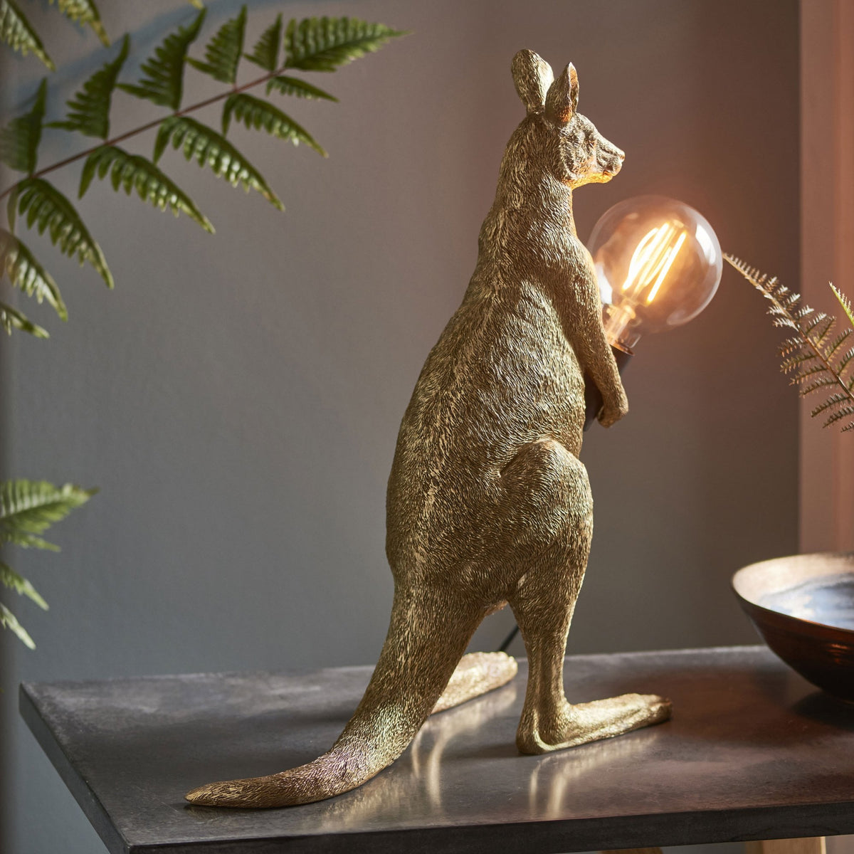 Golden kangaroo table lamp holding a glowing bulb, with green foliage in the background.