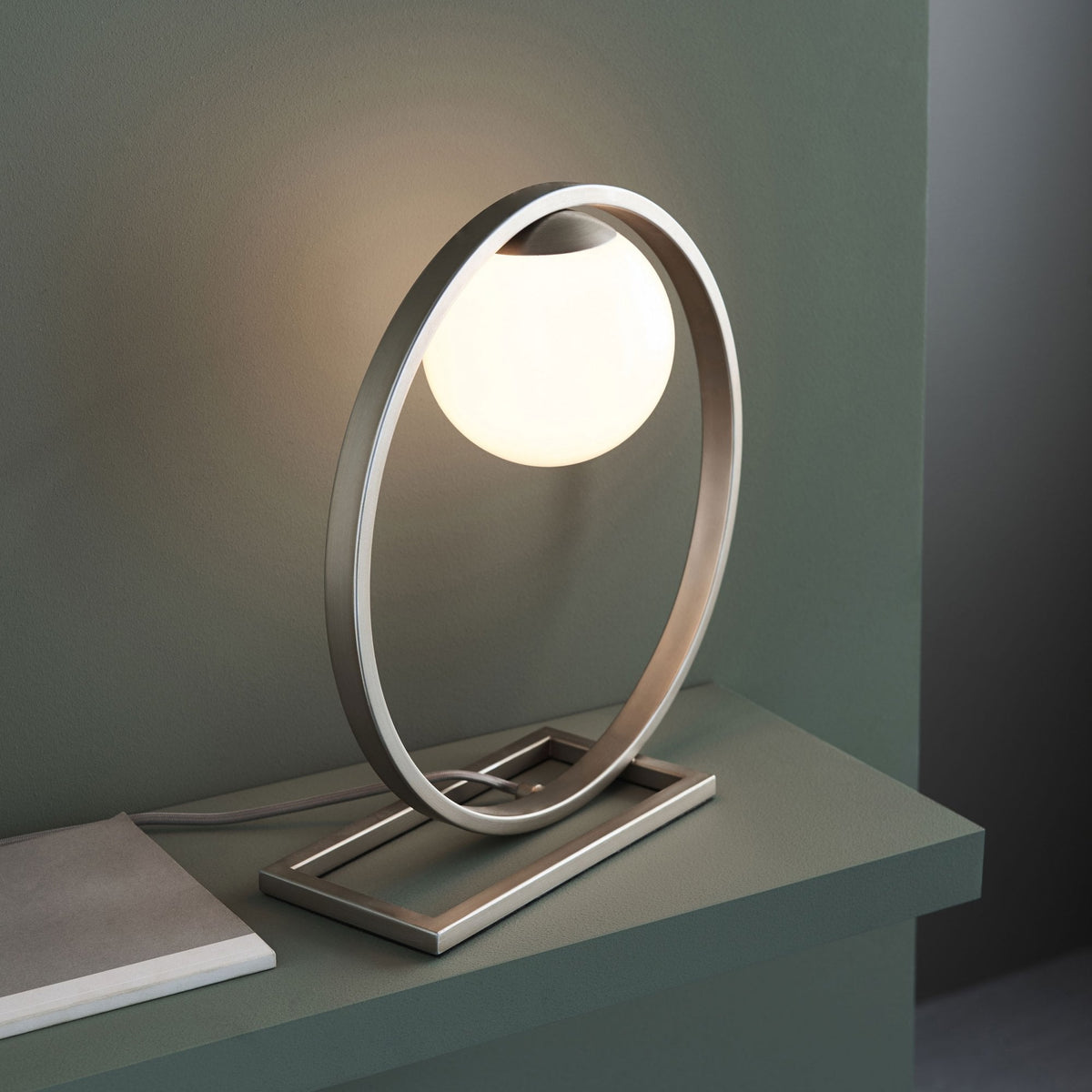 Modern table lamp with a glowing spherical bulb inside a brushed metal oval frame, on a dark green surface.