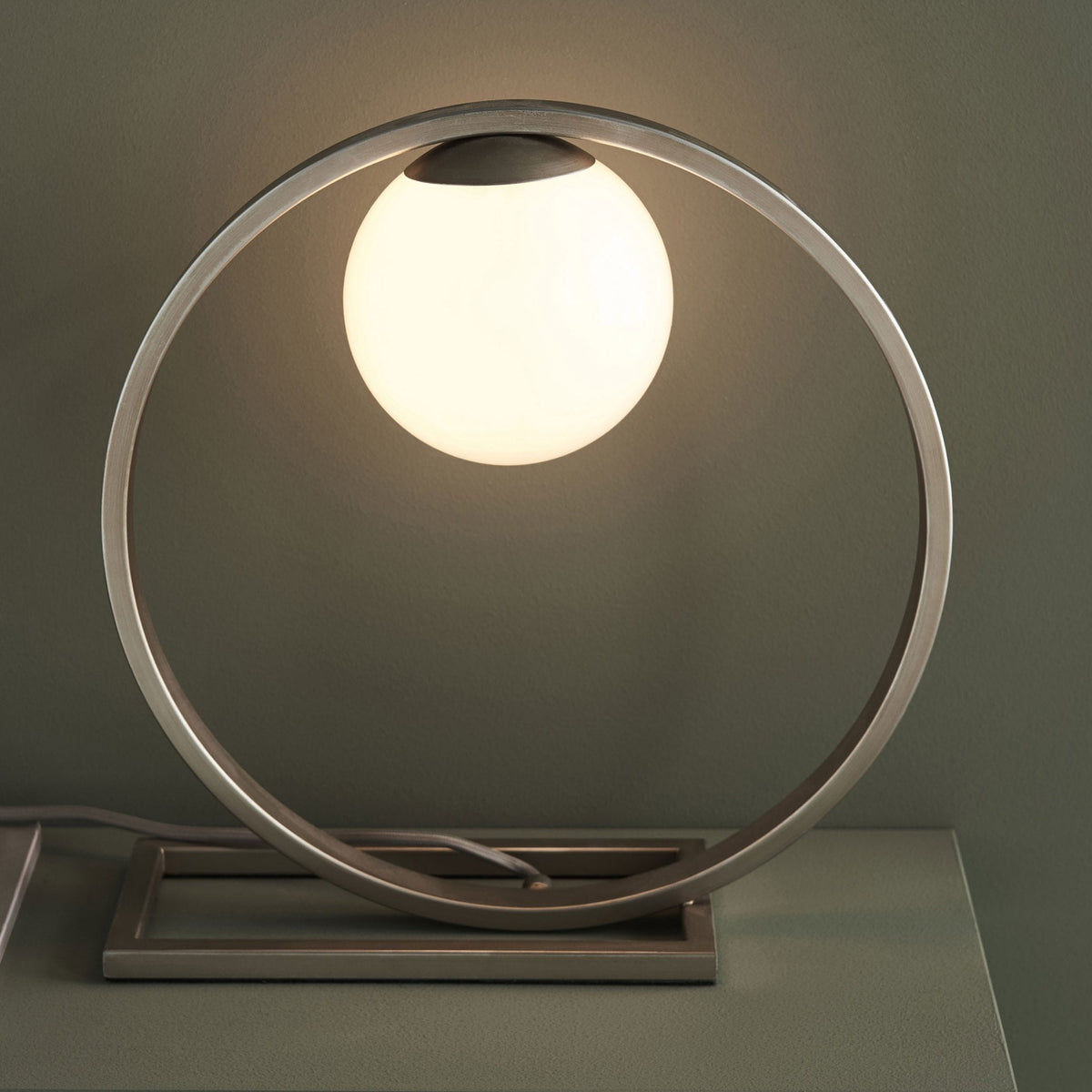 Close-up of a modern table lamp with a glowing spherical bulb inside a brushed metal oval frame.