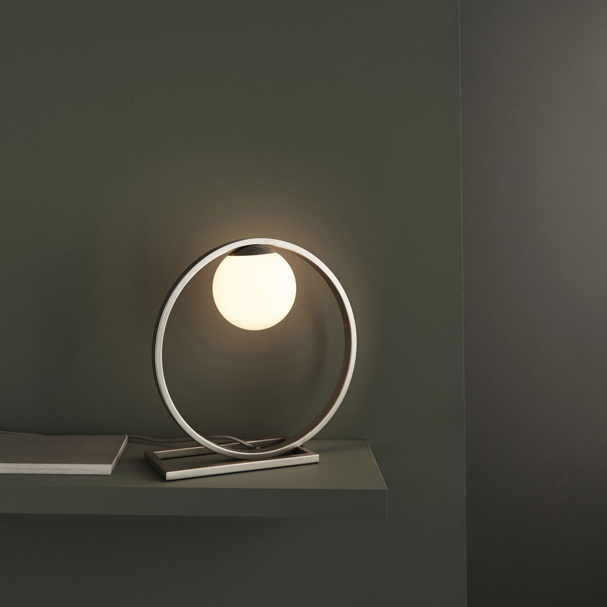 Modern table lamp with a glowing spherical bulb inside a brushed metal oval frame, on a dark green shelf.